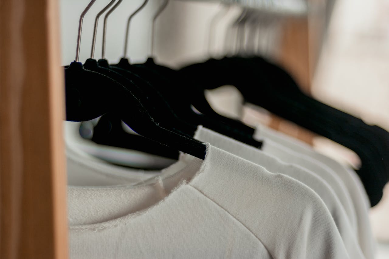 heros-img Close-up of white t-shirts hanging neatly on black hangers in a minimalist indoor closet setting.